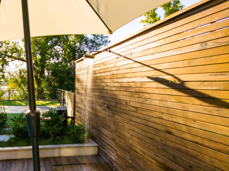 Warmwasser Außendusche mit senkrecht montierter Holzverkleidung, Wasser läuft aus einer schmalen Duscharmatur entlang der Holzwand, im Vordergrund Schirm, im Hintergrund Garten mit Grill.