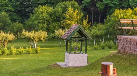 Alter gemauerter Ziehbrunnen mit Holzdach in einem grünen Garten mit Obstbäumen und Sitzbank.