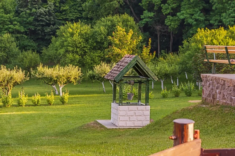Alter gemauerter Ziehbrunnen mit Holzdach in einem grünen Garten mit Obstbäumen und Sitzbank.