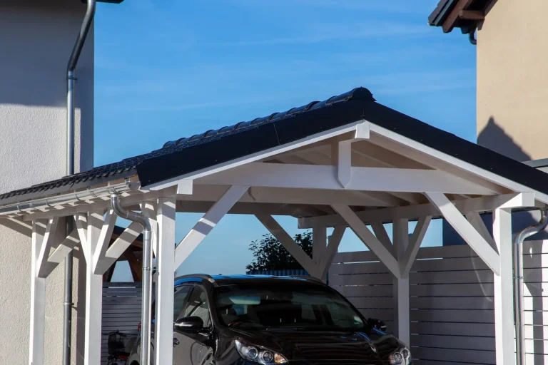 Moderner Carport aus Holz mit Satteldach neben einem Wohnhaus, bietet Schutz für ein geparktes Auto.