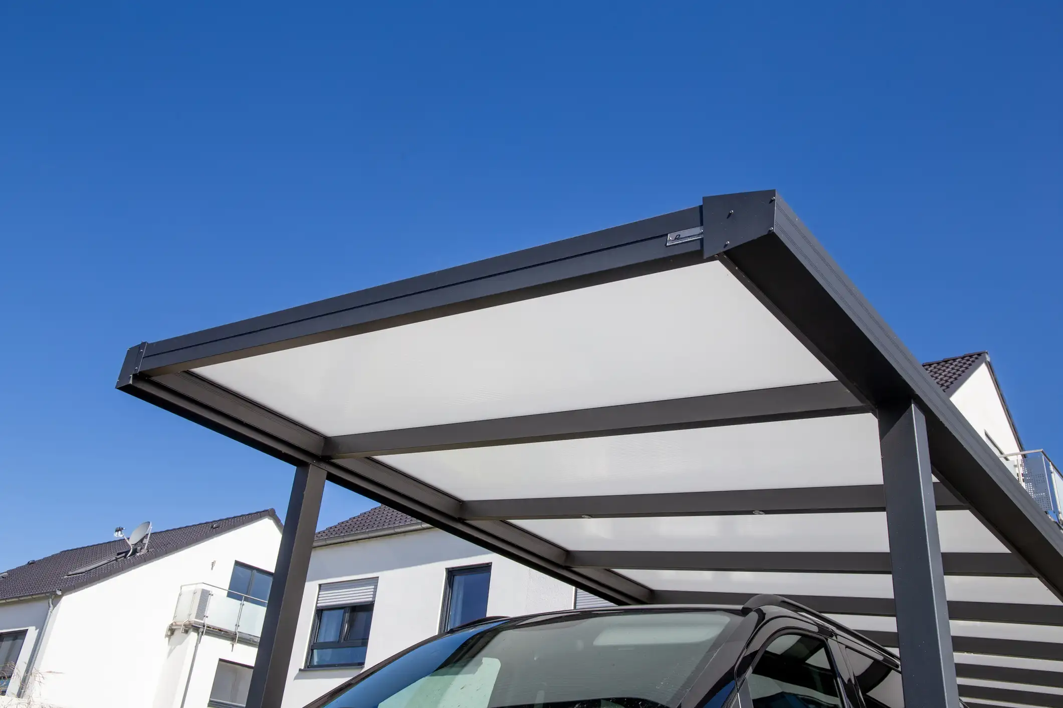 Modernes Carport mit dunklem Metallrahmen und heller Dachbedeckung aus Polycarbonat vor blauem Himmel.
