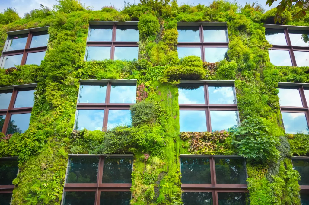 Mehrstöckiges Gebäude mit großen Fensterfronten und dichter wandgebundener Fassadenbegrünung. Unterschiedliche Pflanzenarten wie Moose, Gräser und Sträucher bedecken die Fassade lückenlos. Fokus auf die vertikale Begrünung als nachhaltige Bauweise zur Verbesserung des Stadtklimas.