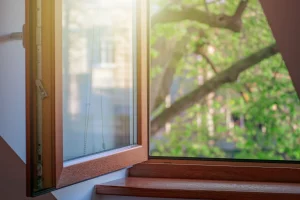 Geöffnetes Holzfenster mit Doppelverglasung, braunem Rahmen und Fensterbank aus Holz, Blick ins Grüne mit Bäumen.