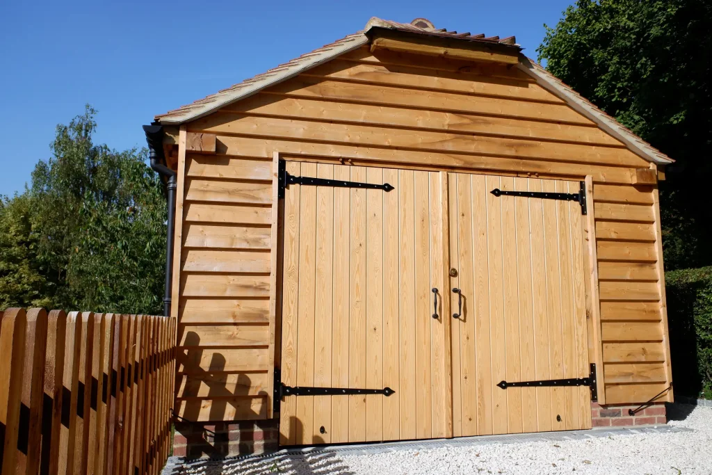 Holzgarage mit Doppeltür und schwarzem Metallbeschlag, neu gebaut aus hellem Holz. Im Vordergrund steht ein Holzzaun, im Hintergrund sind Bäume und blauer Himmel zu sehen.
