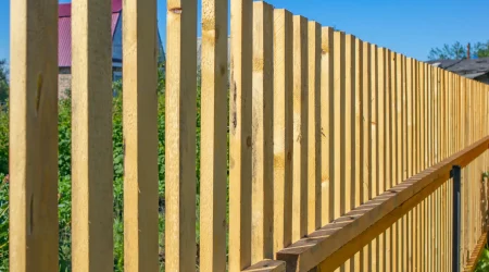 Moderner Holzzaun aus senkrechten Holzlatten mit Metallverstärkung, aufgenommen bei blauem Himmel.