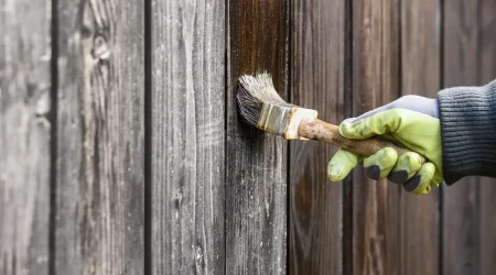 Hand mit Handschuh streicht einen alten Holzzaun mit Holzlasur, um ihn zu reparieren und vor Witterung zu schützen.