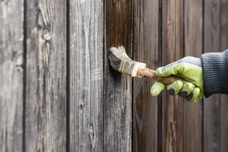 Hand mit Handschuh streicht einen alten Holzzaun mit Holzlasur, um ihn zu reparieren und vor Witterung zu schützen.