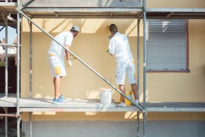 Zwei Handwerker tragen Streichputz auf eine gelbe Hausfassade auf, während sie auf einem Baugerüst arbeiten.