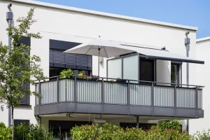 Vorstellbalkon mit grauem Metallgeländer, Markise und Sonnenschirm an einem modernen Wohnhaus mit heller Fassade.