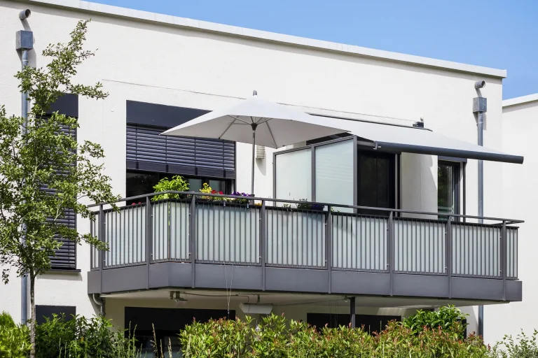 Vorstellbalkon mit grauem Metallgeländer, Markise und Sonnenschirm an einem modernen Wohnhaus mit heller Fassade.