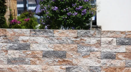 Niedrige Gartenmauer aus rechteckigen, unterschiedlich gefärbten Natursteinblöcken in Grau-, Beige- und Brauntönen, im Hintergrund blühende Sträucher und Bäume in einem gepflegten Garten.