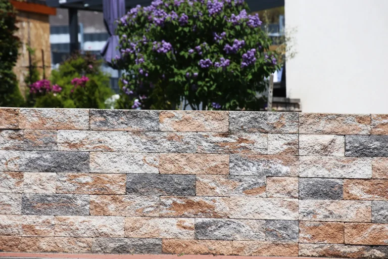 Niedrige Gartenmauer aus rechteckigen, unterschiedlich gefärbten Natursteinblöcken in Grau-, Beige- und Brauntönen, im Hintergrund blühende Sträucher und Bäume in einem gepflegten Garten.