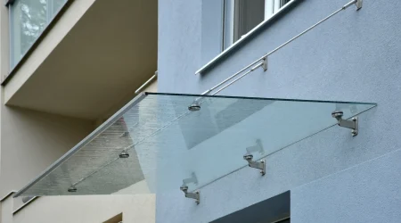 Modernes Glasvordach mit Edelstahlhaltern und Spannseilen an einer Hausfassade, das den Eingangsbereich vor Regen schützt.