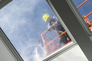 rbeiter mit Schutzhelm reinigt ein Glasdach mit einem Hochdruckreiniger von außen, während Wasser über die Glasfläche läuft.