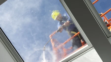 rbeiter mit Schutzhelm reinigt ein Glasdach mit einem Hochdruckreiniger von außen, während Wasser über die Glasfläche läuft.