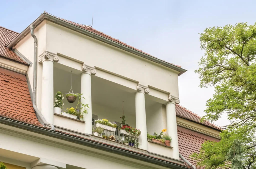 Klassische Loggia mit Säulen und Blumenschmuck an einem Wohnhaus mit rotem Ziegeldach und hellem Putz.
