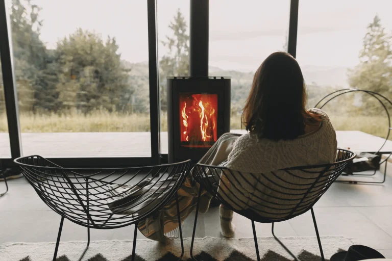 Eine Person sitzt gemütlich vor einem raumluftunabhängigen Kamin in einem modernen Wohnzimmer mit großen Panoramafenstern und blickt auf die Natur draußen.