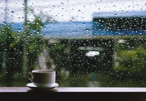 Eine Kaffeetasse steht auf einer Fensterbank vor einer mit Regentropfen bedeckten Scheibe, die Wasser abperlen lässt und die Wirkung von selbstreinigendem Glas zeigt.