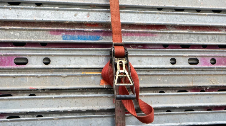 Ein orangener Spanngurt um einen Stapel von Gerüstbauteilen.