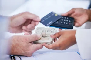 Ein Kunde bezahlt im Fachgeschäft für Hörgeräte. Im Vordergrund werden Hörgeräte und Batterien übergeben. Das Bild veranschaulicht die Kostenübernahme von Hörgeräten durch die Krankenkasse sowie mögliche Zuzahlungen beim Hörgerätekauf.