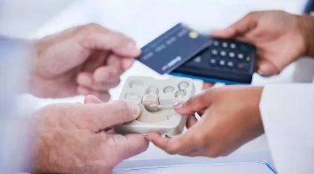 Ein Kunde bezahlt im Fachgeschäft für Hörgeräte. Im Vordergrund werden Hörgeräte und Batterien übergeben. Das Bild veranschaulicht die Kostenübernahme von Hörgeräten durch die Krankenkasse sowie mögliche Zuzahlungen beim Hörgerätekauf.