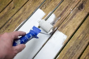 Eine Hand trägt mit einer kleinen Farbrolle weißen Lack auf verwitterte Holzbretter auf.