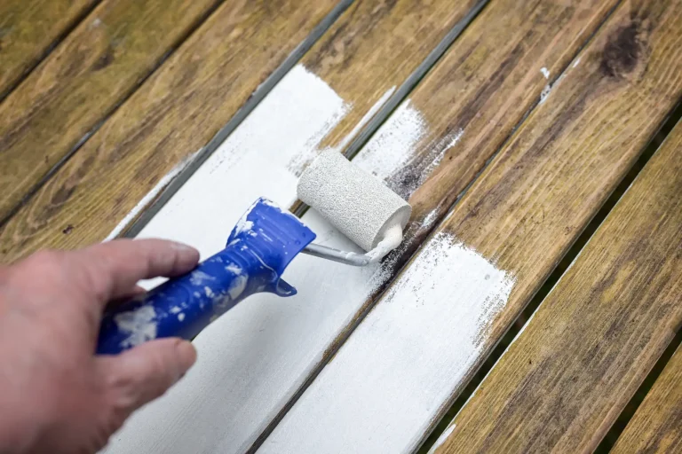 Eine Hand trägt mit einer kleinen Farbrolle weißen Lack auf verwitterte Holzbretter auf.