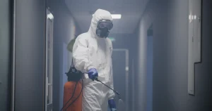 Ein Tatortreiniger in Schutzanzug, Atemmaske und Handschuhen führt in einem Gebäude im Rahmen einer professionellen Tatortreinigung eine Luftdesinfektion durch.