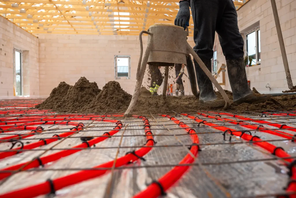 Ein Arbeiter verteilt frischen Estrich über einer Fußbodenheizung in einem Rohbau, um die Restfeuchtigkeit nach Estrichart und die richtige Trocknungsphase zu berücksichtigen.