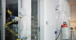 Zwei Tatortreiniger in Schutzanzügen und Atemmasken entfernen Absperrband und desinfizieren einen Raum nach einem Polizeieinsatz.