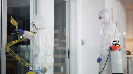 Zwei Tatortreiniger in Schutzanzügen und Atemmasken entfernen Absperrband und desinfizieren einen Raum nach einem Polizeieinsatz.