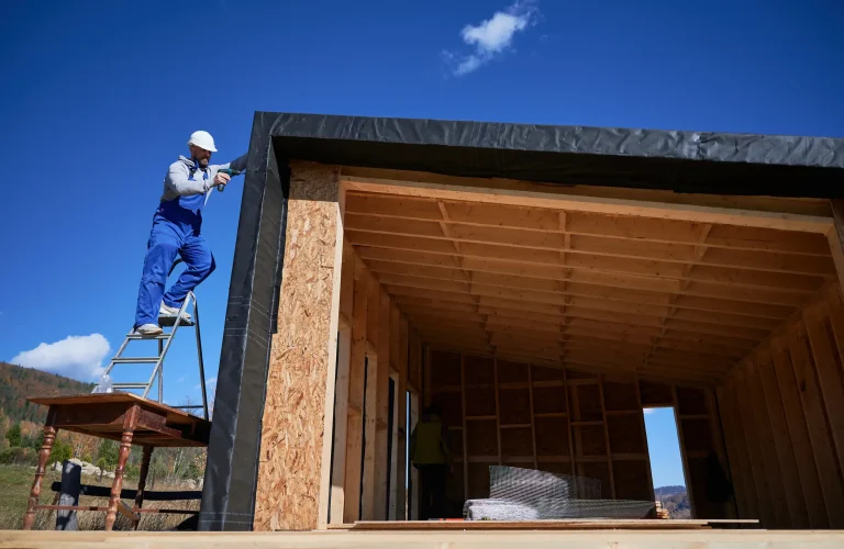 Handwerker steht auf einer Leiter und montiert eine Dampfsperrfolie am Dach eines Tiny Houses. Im Inneren ist die Holzkonstruktion und die noch unvollständige Dämmung zu sehen.