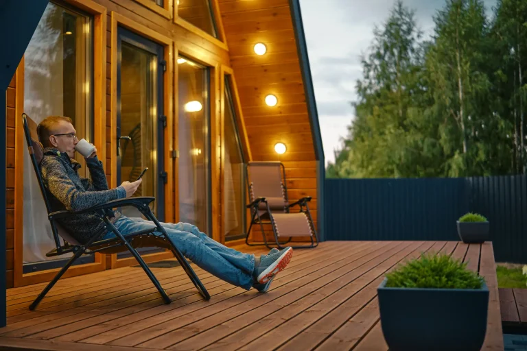 Ein Mann sitzt entspannt auf der Holzterrasse eines modernen Tiny Houses, trinkt Kaffee und schaut auf sein Smartphone, während im Hintergrund warme Außenbeleuchtung und Bäume zu sehen sind.