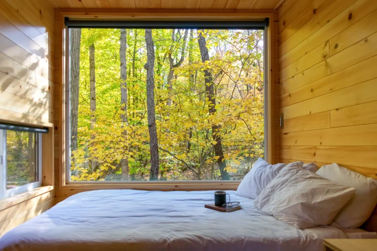 Großes Fenster in einem Tiny House mit Holzverkleidung, Blick auf einen herbstlichen Wald mit gelben Blättern.