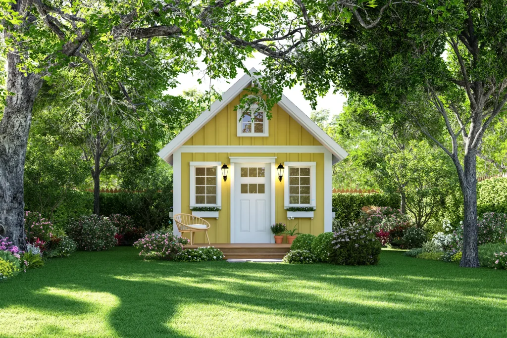 Kleines gelbes Tiny House mit weißen Sprossenfenstern, Giebel und Vorgarten mit Blumen und Rasen.