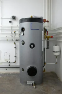 Zylindrischer Warmwasserspeicher in einem Hauswirtschaftsraum, umgeben von mehreren Wasserleitungen und Armaturen.