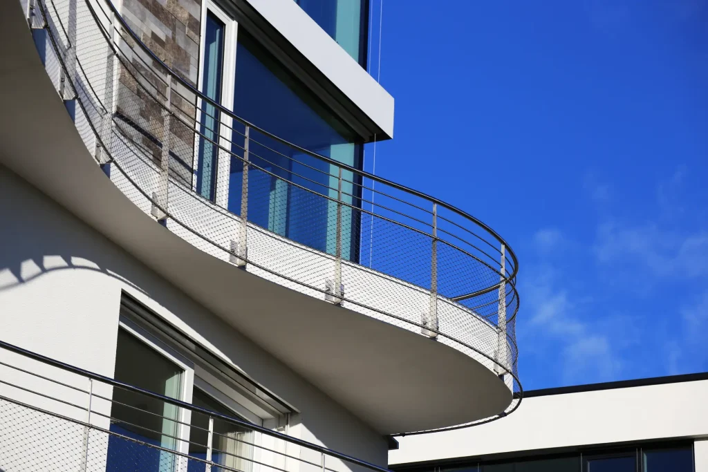 Ein modernes Gebäude mit einem geschwungenen Balkon. Das Balkongeländer aus Edelstahl besteht aus senkrechten Pfosten, horizontalen Streben und einem feinmaschigen Metallnetz. Hinter dem Balkongeländer aus Edelstahl sind große Glasfenster und eine helle Fassadenfläche sichtbar. Der Balkon verläuft entlang der Gebäudefront vor blauem Himmel.