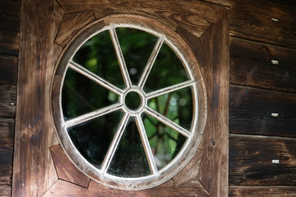 Ein rundes Holzfenster mit mehreren Glasfeldern ist in eine dunkle Holzwand eingelassen. Durch die Glasscheiben sind grüne Pflanzen im Hintergrund erkennbar. Der Holzrahmen weist eine natürliche Maserung und eine rustikale Oberfläche auf.