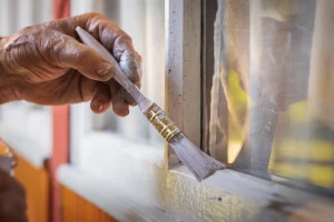 Eine Hand hält einen Pinsel und trägt eine weiße Beschichtung auf den unteren Rahmen eines Fensters auf. Der Fensterrahmen besteht aus Holz und grenzt an eine Glasscheibe. Die Oberfläche des Rahmens ist teilweise bereits gestrichen, während angrenzende Bereiche noch unbehandelt sind.