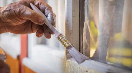 Eine Hand hält einen Pinsel und trägt eine weiße Beschichtung auf den unteren Rahmen eines Fensters auf. Der Fensterrahmen besteht aus Holz und grenzt an eine Glasscheibe. Die Oberfläche des Rahmens ist teilweise bereits gestrichen, während angrenzende Bereiche noch unbehandelt sind.