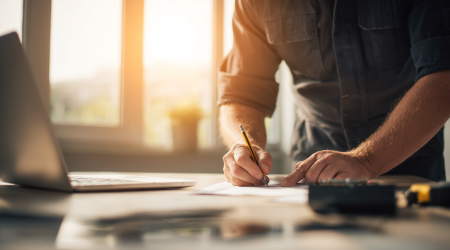 Ein Elektriker in Arbeitskleidung repariert eine Steckdose, während im Vordergrund ein Schreibtisch mit Laptop, PDF-Rechnung und Stift zu sehen ist, der die Verbindung von handwerklicher Arbeit und Büroaufgaben zeigt.