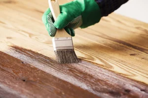 Eine Person trägt grüne Arbeitshandschuhe und trägt mit einem Pinsel Holzlasur auf eine Holzoberfläche auf. Die Lasur wird gleichmäßig entlang der Holzmaserung aufgetragen und dunkelt das Holz sichtbar nach.