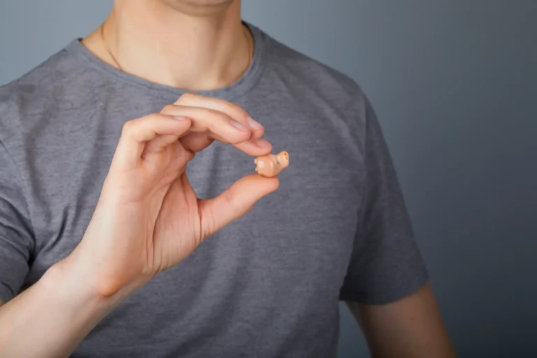 Nahaufnahme einer Person im grauen T-Shirt, die ein ITC-Hörgerät zwischen Daumen und Zeigefinger hält. Das beigefarbene In-the-Canal-Hörgerät ist klein und für den Teil des Gehörgangs konzipiert. Der Hintergrund ist neutral, sodass der Fokus auf dem Hörgerät liegt.