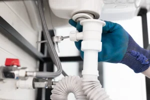 Eine Person mit Arbeitshandschuhen überprüft den Anschluss des Siphons unter einem Waschbecken. Sichtbar sind der weiße Kunststoffsiphon, ein flexibler Abwasserschlauch und ein Anschlussventil mit rotem Griff.