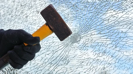 Hand mit Hammer schlägt auf ein Fensterglas, das stark splittert.
