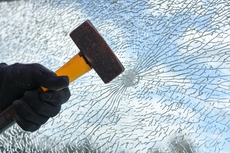 Hand mit Hammer schlägt auf ein Fensterglas, das stark splittert.