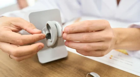Eine Akustikerin hält ein Hörgerät in beiden Händen und setzt ein Ohrpassstück in das graue Hörgerätgehäuse ein. Das Hörgerät liegt vor einer weißen Halterung auf einem Holztisch.