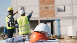 Zwei Bauarbeiter auf einer Baustelle, besprechen das weitere Vorgehen am Rohbau. Im Vordergrund liegen zwei Schutzhelme auf einem Tisch.