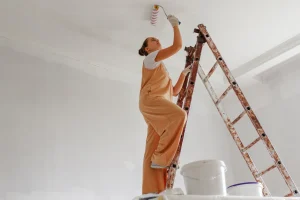 Frau in Malerkleidung streicht weiße Decke mithilfe einer Leiter.