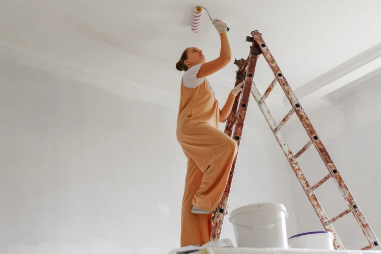Frau in Malerkleidung streicht weiße Decke mithilfe einer Leiter.